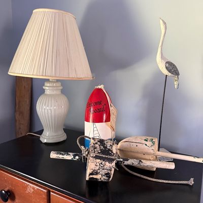 Sale Photo Thumbnail #22: Nautical decor arrangement on a dark tabletop featuring a white ceramic lamp with a pleated shade, a red and white decorative lobster buoy, a carved wooden heron sculpture, and vintage painted fishing floats.