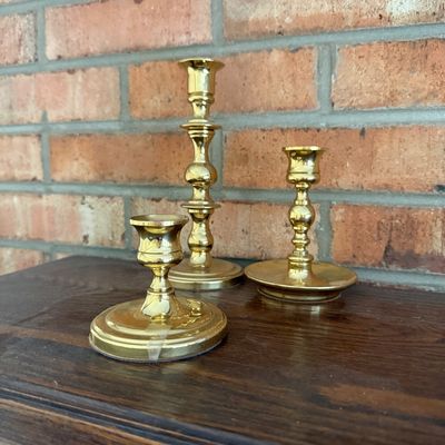 Sale Photo Thumbnail #50: Set of three polished brass candlesticks in varying heights, displayed on a wooden surface against a brick wall.