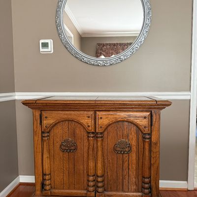 Sale Photo Thumbnail #83: Traditional oak server or cabinet with arched panel doors and decorative hardware, paired with a round silver-framed wall mirror.