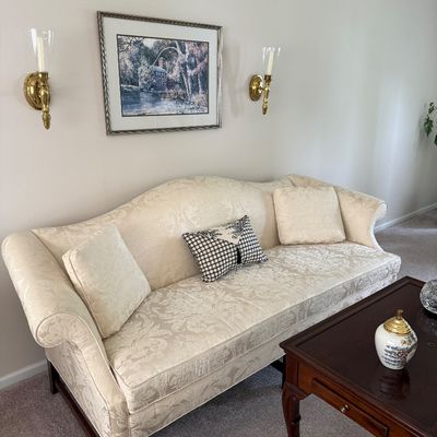 Sale Photo Thumbnail #76: Traditional cream damask upholstered sofa with camelback and rolled arms, flanked by a pair of brass wall sconces and framed landscape artwork above.