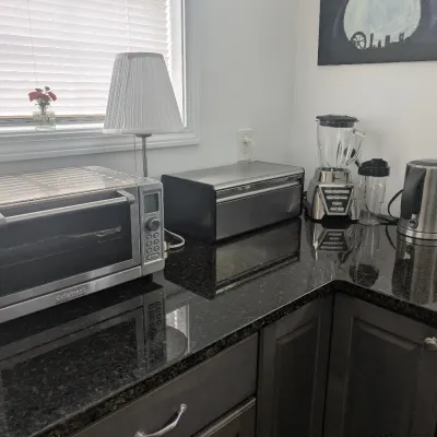Collection of modern stainless steel kitchen counter appliances, including a Cuisinart toaster oven, bread box, Oster blender, and...