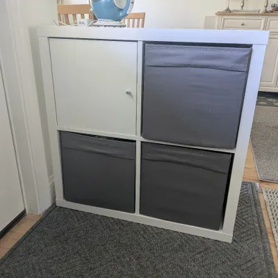 White 2x2 IKEA Kallax style cube shelving unit, featuring one cabinet insert door and three gray fabric storage bins.