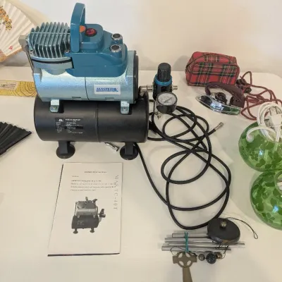 Master Airbrush TC-40T air compressor with tank and manual, displayed alongside vintage folding fans, a travel iron, wind chimes, and...