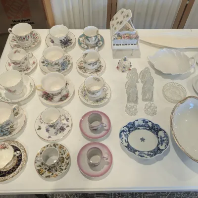 Collection of assorted fine china teacups, saucers, serving dishes, and a decorative miniature piano figurine arranged on a white table.
