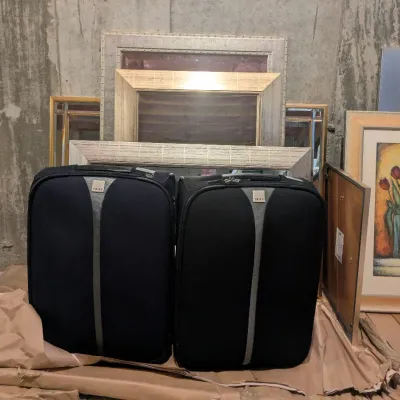 Pair of black Tripp rolling carry-on suitcases alongside a collection of framed artwork and empty picture frames against a concrete wall.