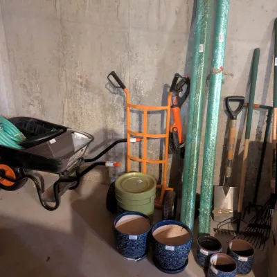 Collection of yard work tools including a black wheelbarrow, an orange hand truck, long-handled shovels, rakes, ceramic planters, and...