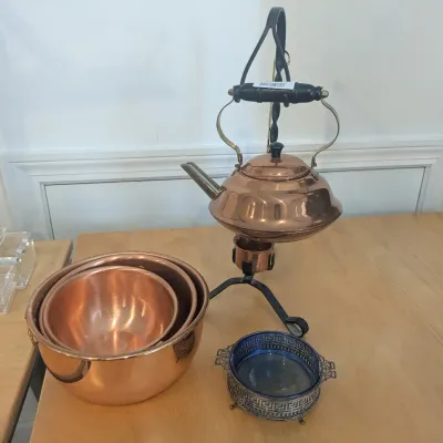 Vintage copper tilt kettle on a black wrought iron burner stand, displayed alongside a set of nested copper mixing bowls and a small...