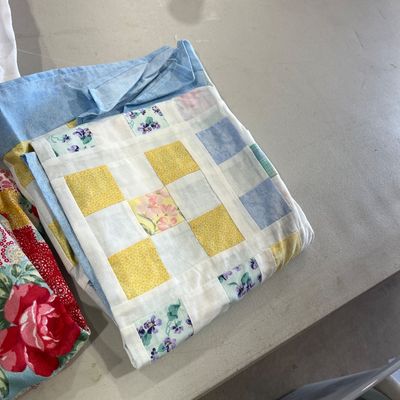 Sale Photo Thumbnail #121: Folded handmade patchwork quilt with yellow, blue, and floral fabrics on a table alongside other textiles.