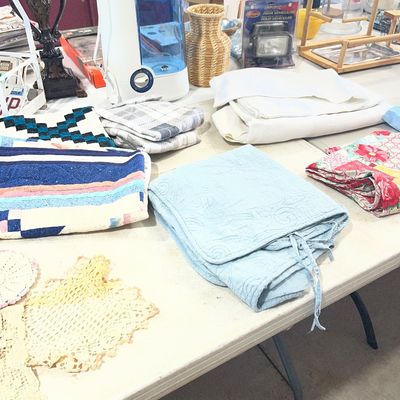 Sale Photo Thumbnail #116: A table arranged with household items including folded quilts, linens, crocheted doilies, a Vicks humidifier, and a small woven vase.