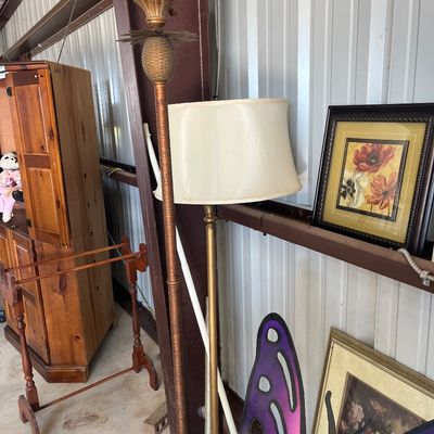 Sale Photo Thumbnail #51: A brass floor lamp with a cream shade displayed alongside a decorative pineapple-topped lamp pole, framed floral artwork, and a tall wooden cabinet.
