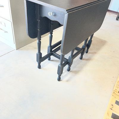 Sale Photo Thumbnail #128: Black painted drop-leaf side table with turned legs, a single drawer featuring a clear glass knob, and a distressed finish.