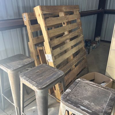 Sale Photo Thumbnail #89: Set of three industrial metal bar stools with distressed tops, shown alongside wooden shipping pallets in a storage area.