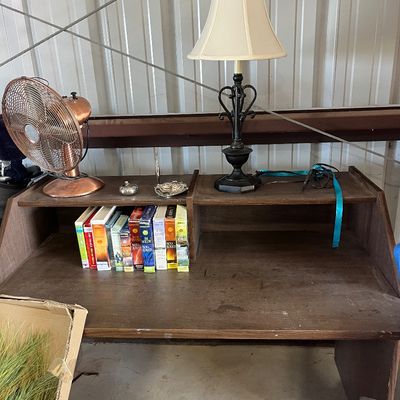 Sale Photo Thumbnail #44: Dark wood student desk with a top shelf featuring an assortment of paperback books, a copper-finish electric fan, and a black wrought-iron table lamp with a beige fabric shade.