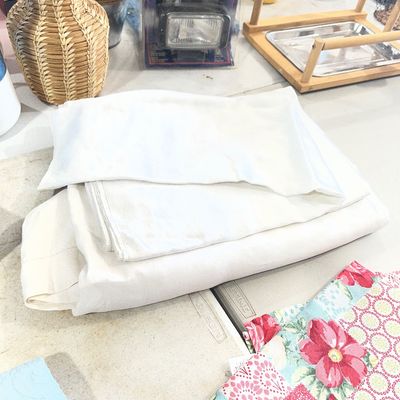 Sale Photo Thumbnail #120: Collection of household goods including folded white linens, a colorful floral quilt, a woven wicker vase, packaged automotive driving lights, and a wooden pet perch.