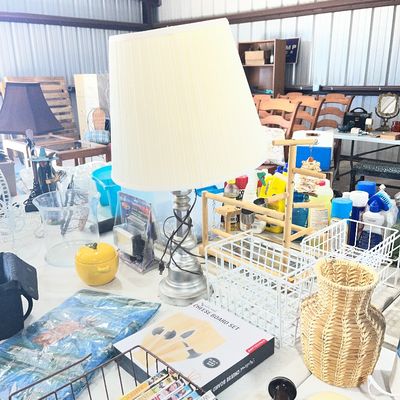 Sale Photo Thumbnail #96: A silver-tone urn-style table lamp with a large pleated fabric shade, surrounded by various household goods including wire storage baskets, DVDs, and a woven vase.