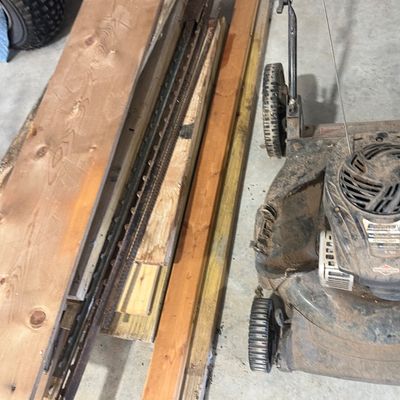 Sale Photo Thumbnail #17: A stack of assorted lumber, wood boards, and metal tracks alongside a dusty Bolens walk-behind lawn mower on a concrete floor.