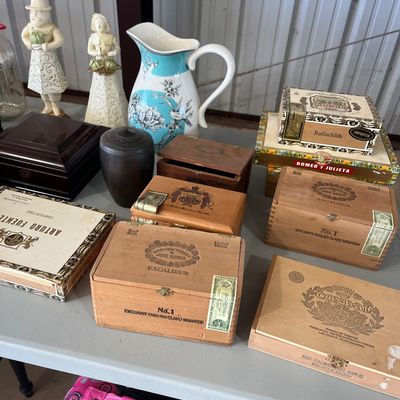 Sale Photo Thumbnail #10: Collection of vintage wooden and paper cigar boxes, displayed alongside decorative figurines, a floral ceramic pitcher, and a dark urn.