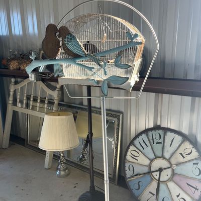 Sale Photo Thumbnail #122: Vintage-style birdcage on a metal floor stand, displayed alongside a decorative lovebirds sculpture, large oversized wall clock, and table lamp.
