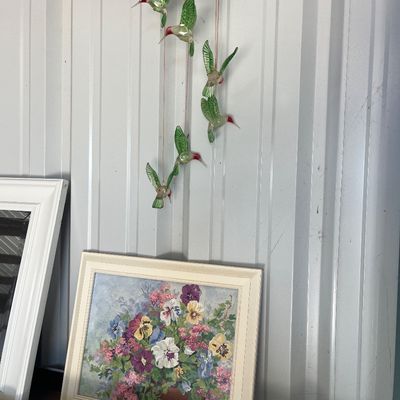 Sale Photo Thumbnail #62: Framed floral painting featuring colorful pansies, sitting below a hanging decorative solar-powered hummingbird wind chime sculpture.