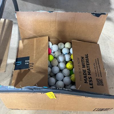 Sale Photo Thumbnail #39: Cardboard Amazon box filled with a large quantity of assorted golf balls in white, yellow, and pink.