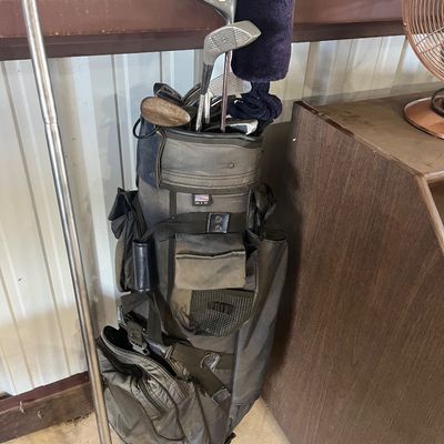 Sale Photo Thumbnail #83: Vintage dark gray cart golf bag filled with assorted golf clubs and driver covers, showing signs of wear.