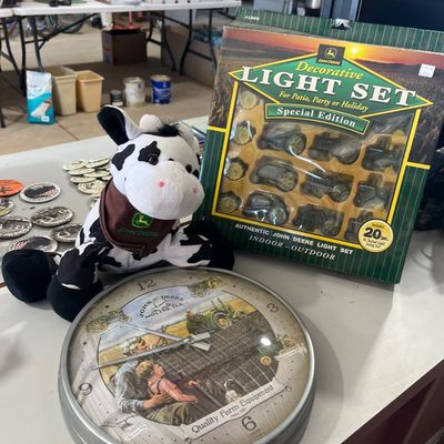 Sale Photo Thumbnail #107: John Deere collectibles including a round wall clock featuring vintage farm artwork, a plush cow wearing a John Deere bandanna, and a boxed 20-piece decorative light set.