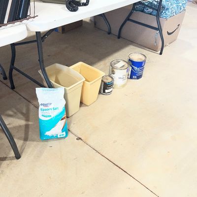 Sale Photo Thumbnail #102: Assorted household supplies including an 8-pound bag of Equate Epsom Salt, plastic wastebaskets, paint cans, and a vintage yellow rotary telephone on a folding table.