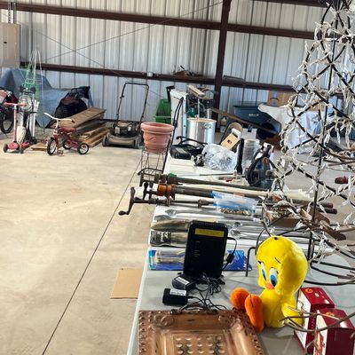 Sale Photo Thumbnail #37: Assorted estate sale items arranged on tables inside a workshop, including a plush Tweety Bird toy, copper tray, hand tools, long bar clamps, and various outdoor equipment in the background.