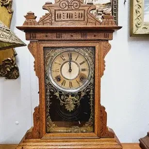 Antique pressed oak mantel clock featuring carved crest details, Roman numerals, and a gold-etched glass door panel.