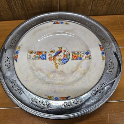 Vintage ceramic cake plate featuring a colorful bird and floral design, resting in a reticulated metal holder with a swinging handle.