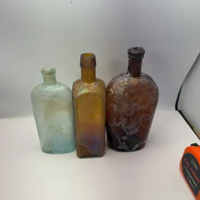Trio of antique glass bottles, including an aqua flask, an amber square bottle, and a dark amber flask, all displaying natural...