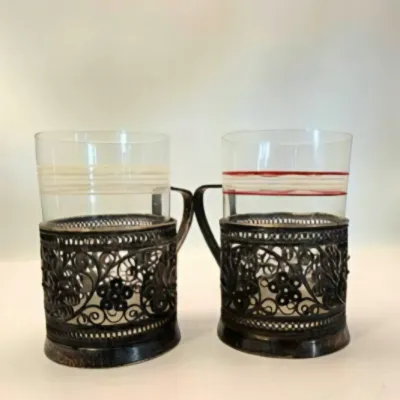 Pair of Russian style podstakannik tea glasses featuring ornate filigree metal holders and thin glass inserts with striped detailing.