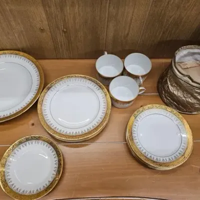 Vintage white porcelain dinnerware set with ornate gold filigree rims, including plates, bowls, teacups, and a quilted storage bag.