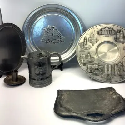 Collection of pewter tableware items, including decorative plates featuring a sailing ship and German landmarks, a White Horse Scotch...