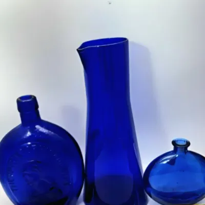 Trio of vintage cobalt blue glassware pieces, including an embossed flask-style bottle, a tall pitcher with a pour spout, and a small...