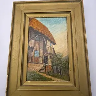 Framed original oil painting depicting a rustic thatched-roof cottage with half-timbered walls, set in a wide molded gold-painted wooden...