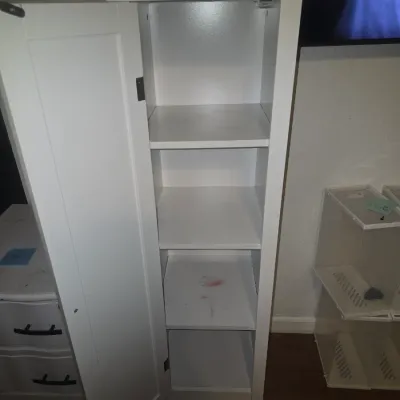 Sale Photo Thumbnail #34: A tall, narrow freestanding storage cabinet or pantry in a painted white finish. Features a single door with a beadboard accent panel, round silver knob, crown molding trim at top, and a subtle arched bottom apron. Appears in good overall pre-owned condit