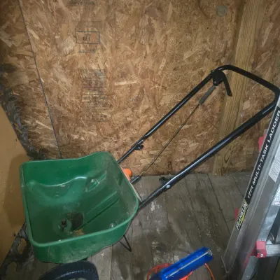 Sale Photo Thumbnail #150: Walk-behind green lawn fertilizer and seed broadcast spreader featuring a plastic hopper, black metal handle, and two wheels. Includes an orange extension cord and a blue metal caulking gun. Items show signs of light use and dirt consistent with normal ya