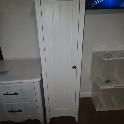 Sale Photo Thumbnail #33: A tall, narrow freestanding storage cabinet or pantry in a painted white finish. Features a single door with a beadboard accent panel, round silver knob, crown molding trim at top, and a subtle arched bottom apron. Appears in good overall pre-owned condit