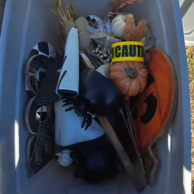 Sale Photo Thumbnail #114: Plastic storage tub containing a variety of Halloween and seasonal decorations. Includes wooden spiderweb cutout signs, a glittery pumpkin sign, artificial florals, a caution tape pumpkin stake, ghost decor, and black cat/cauldron accent pieces. Items app
