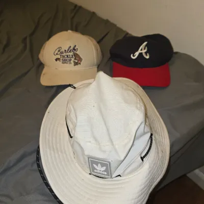 Sale Photo Thumbnail #199: Lot of three men's hats including a white Adidas sun/boonie hat with neck cord, a cream/beige Burlebo Tackle Shop Supply Co. baseball cap with fish graphic, and a navy/red Atlanta Braves style baseball cap with embroidered 'A'. All hats show light pre-own