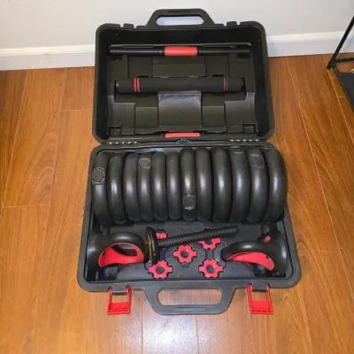 Sale Photo Thumbnail #190: An adjustable weight set including multiple plastic-coated weight plates, dumbbell bars, kettlebell handles, star collar locks, and a connecting rod, all housed in a molded black plastic carrying case with red latches. Appears lightly used in good conditi