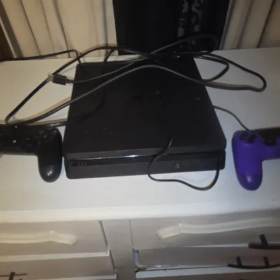 Sale Photo Thumbnail #59: Sony PlayStation 4 (PS4) Slim gaming console in matte black. Includes two DualShock 4 wireless controllers (one black, one purple) and connecting cables. Shows minor surface scuffs and signs of normal wear. Small melt spot