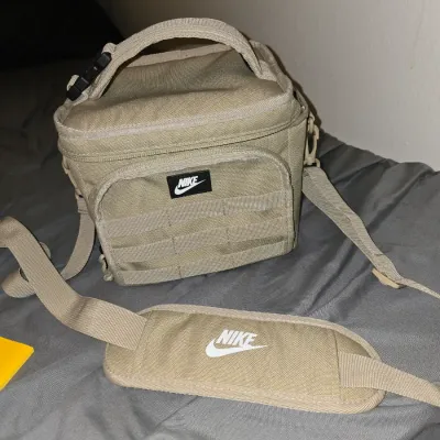 Sale Photo Thumbnail #196: Nike soft-sided insulated lunch bag or mini cooler in a tan/khaki color. Features a top carry handle, a front zippered pocket with MOLLE-style webbing, an adjustable clip top handle, and a removable shoulder strap with a padded Nike branded strap sleeve. 