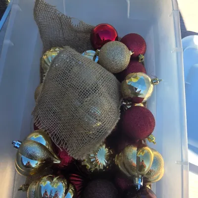 Sale Photo Thumbnail #140: Large white deco mesh Christmas wreath decorated with festive signs ('NOEL', 'JOY', 'PEACE'), faux peppermint candies, and a red bow. Included is a clear plastic storage tote filled with a variety of red, gold, and burgundy spherical and onion-shaped orna