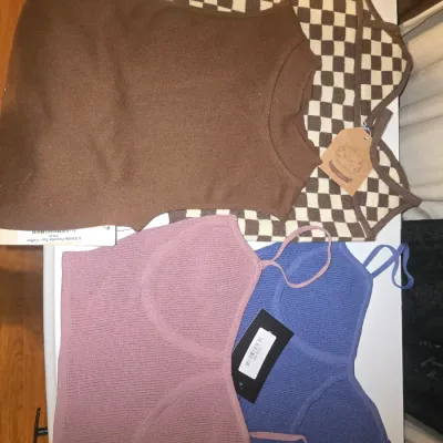 Sale Photo Thumbnail #84: Assorted lot of women's apparel including tank tops, cropped tops, knit sweater vests (including checkered patterns and 'miss love' brand), a pink tiered ruffle top, a white lace-trimmed bodysuit, a gold crop top, and knit socks. Items are lightly used to