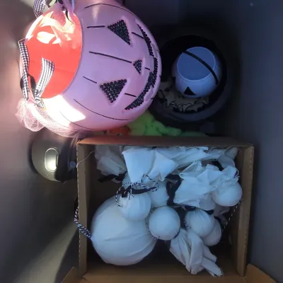 Sale Photo Thumbnail #118: A plastic tote filled with modern Halloween decorations and accessories. Includes a pink jack-o'-lantern bucket with tulle and bow accents, mini cauldrons, decorative fabric ghost favors or ornaments, green faux cobweb fluff, and additional small items. P