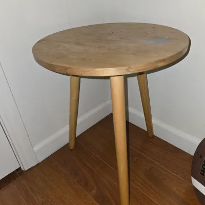 Sale Photo Thumbnail #186: Small round wooden side or accent table featuring three tapered legs in a Mid-Century Modern style design. Shows surface wear, dust, and minor staining/discoloration on the tabletop surface. Overall sturdy construction.