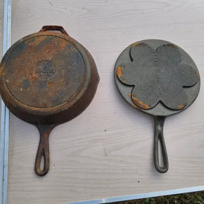 Sale Photo Thumbnail #169: Lot of two cast iron cookware items. Includes a Lodge cast iron skillet with dual pour spouts and a helper handle, alongside a 7-socket silver dollar pancake / Swedish plett pan featuring a flower design on the underside. Both pieces show surface rust and