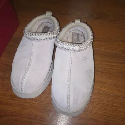 Sale Photo Thumbnail #39: Pair of UGG Tasman style slip-on slippers in light grey suede featuring braided detail along the collar and shearling lining. Shows visible wear, smudges, and discoloration on the suede uppers and interiors from use.